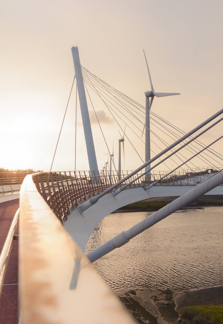 Suspension Bridge Over River And Wind Turbines In Morning Light