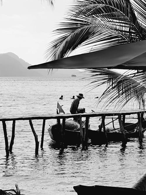 Grayscale Photo of Man Standing on Boat Beside a Boardwalk