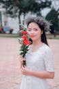 一位穿着白色连衣裙、头戴花冠的亚洲女子，在公园里拿着一支红玫瑰