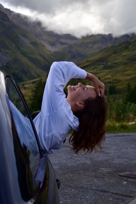 Woman in white laughing, leaning out of car window with stunning Austrian mountain backdrop.