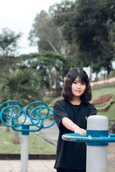 Asian woman exercising in a green park, surrounded by trees and fitness equipment.