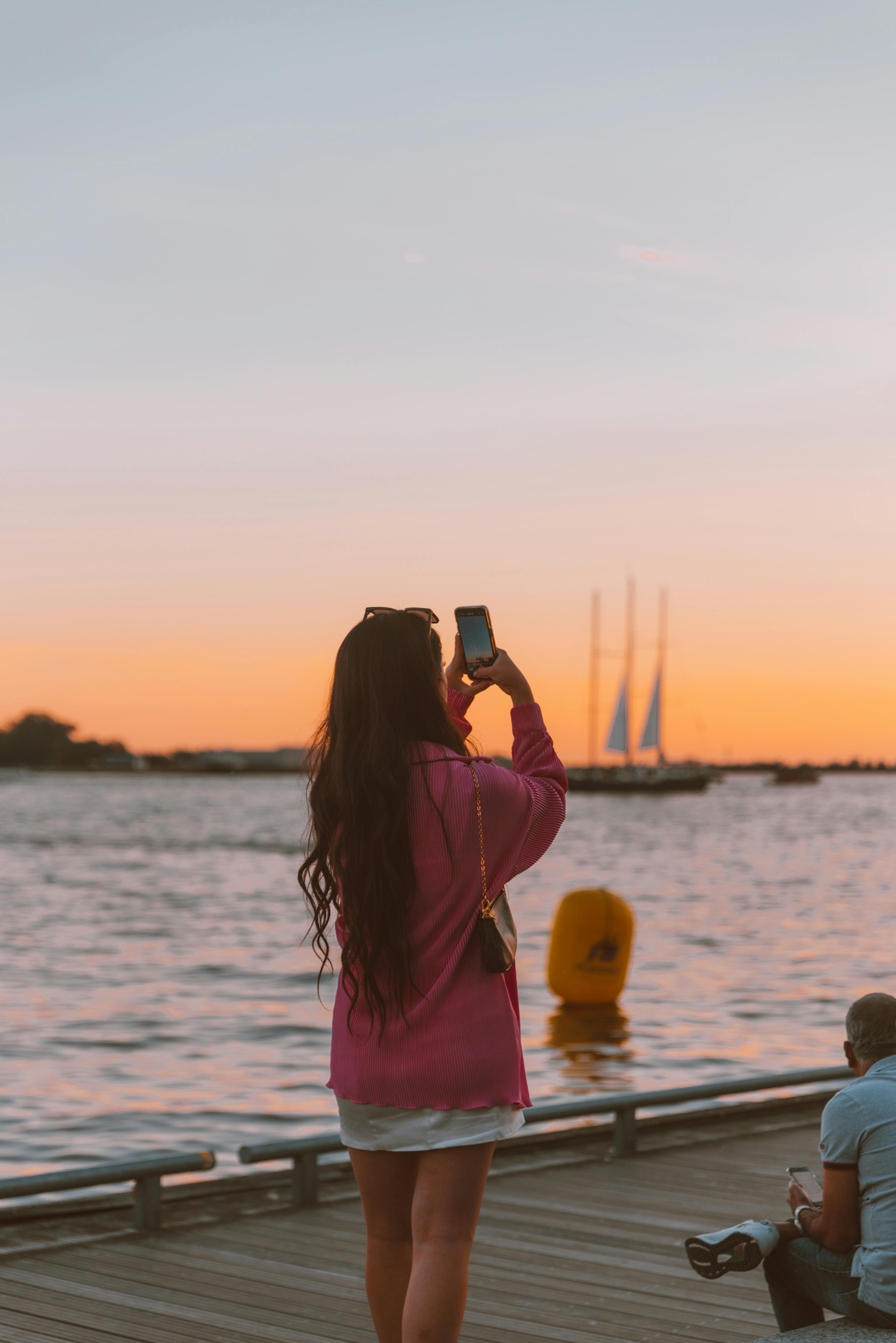 Silhouette of woman capturing sunset over water, vibrant sky.