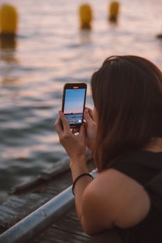 A woman takes a sunset photograph with her smartphone by the seashore. Ideal for lifestyle and travel themes.