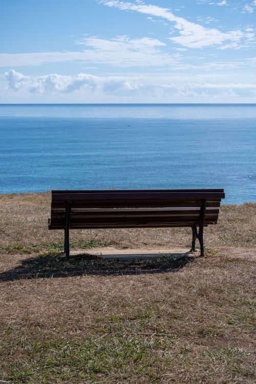Free A solitary bench facing a calm ocean under a bright blue sky, perfect for a serene outdoor escape. Stock Photo