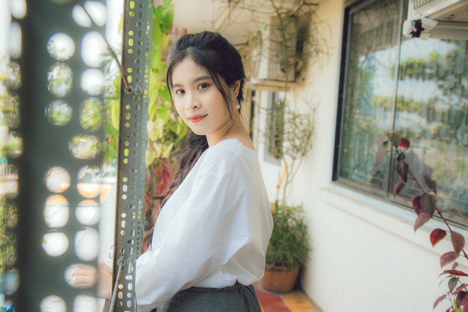 A young woman in white top poses on a bright balcony with plants.