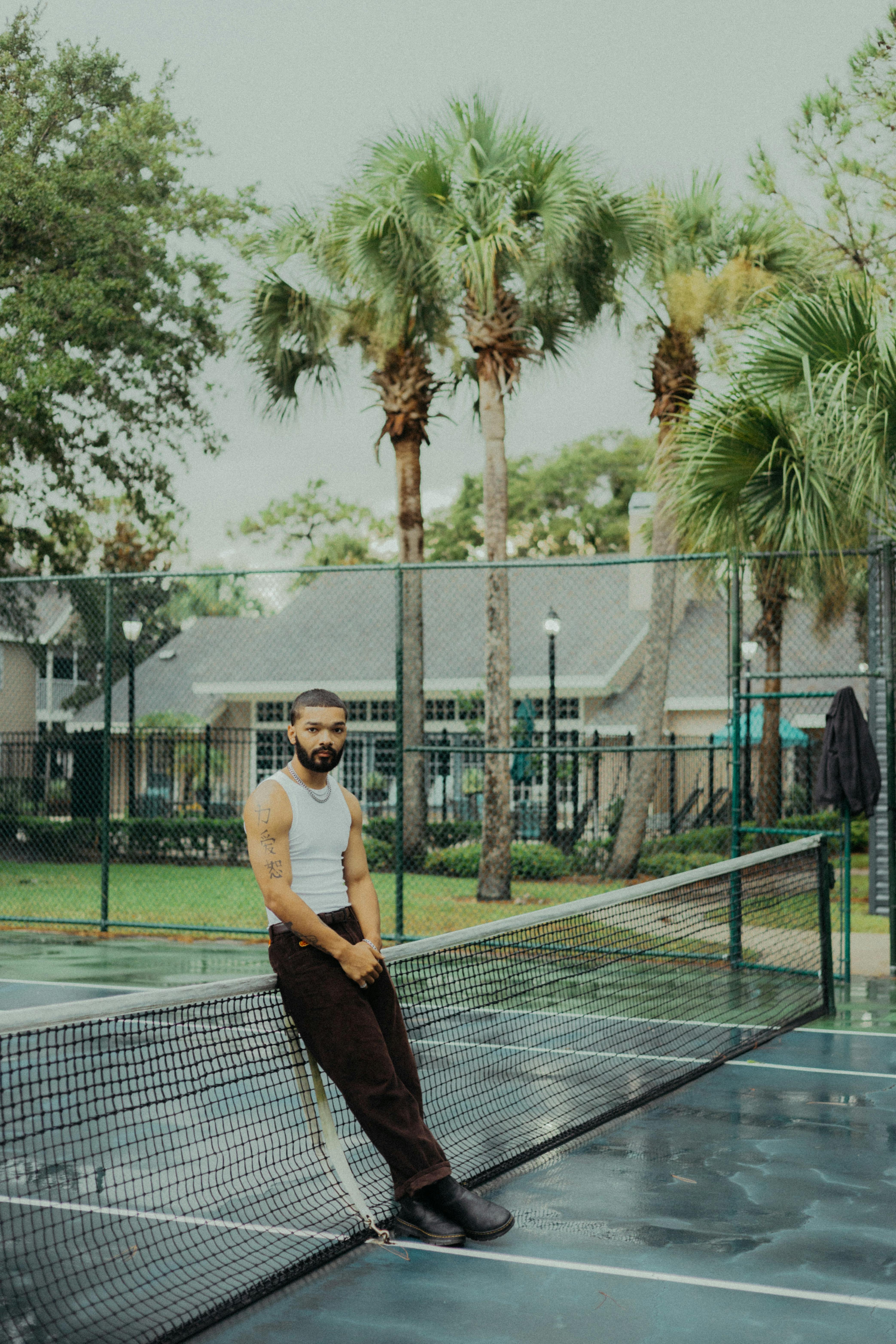 Bearded man leaning on tennis net in a relaxed outdoor setting with palm trees.