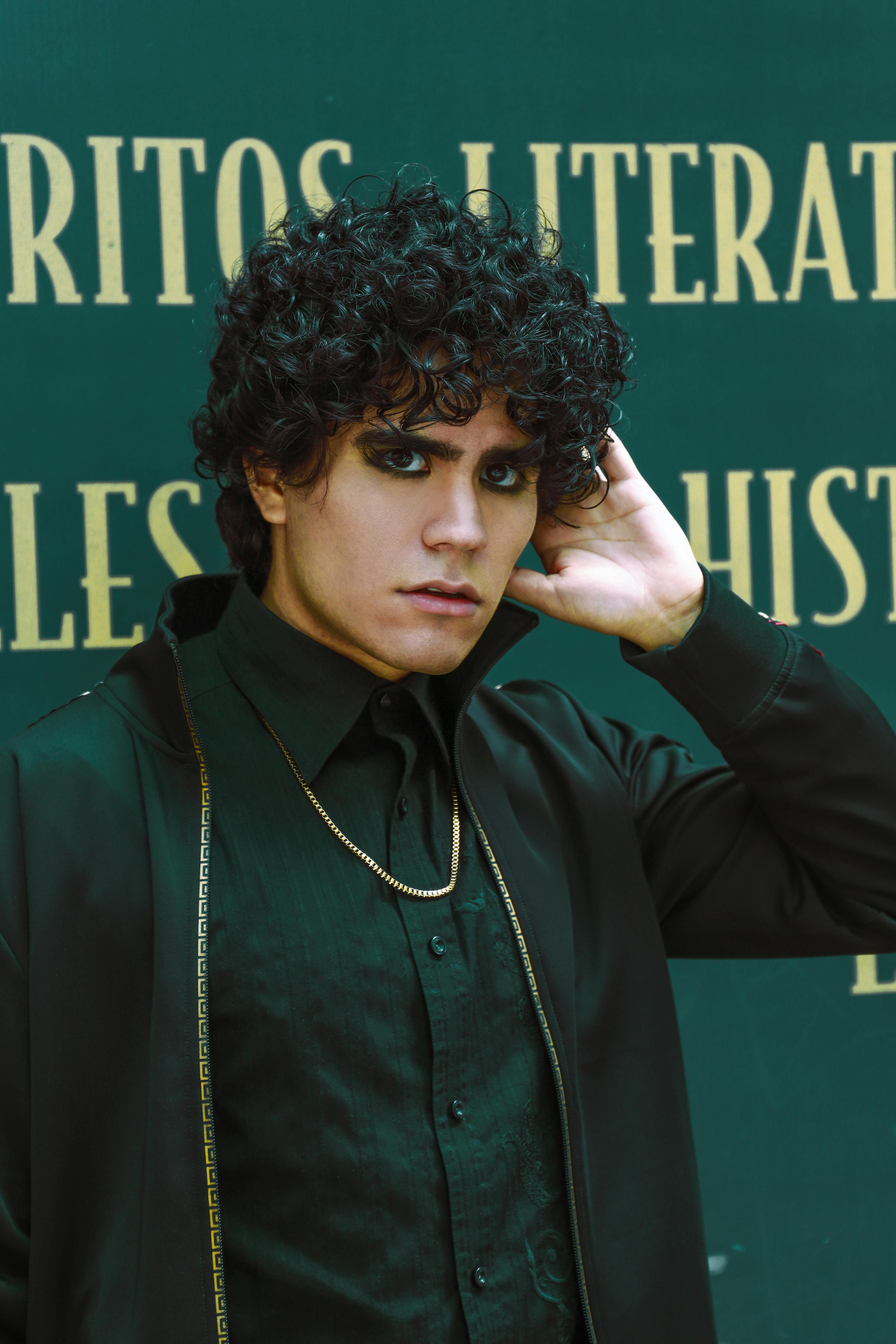 Striking portrait of a man in black attire with curly hair and bold makeup.