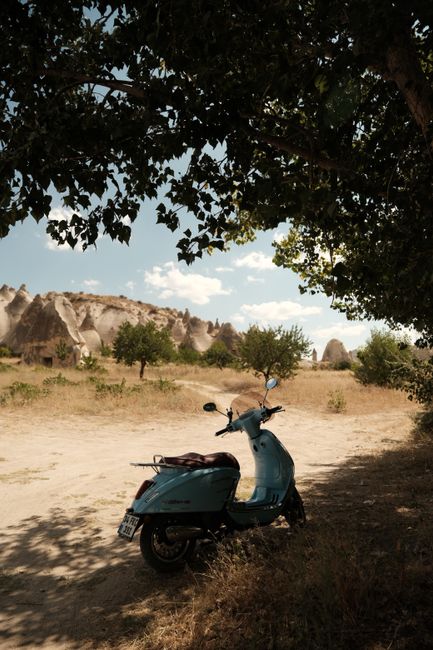 A parked blue scooter under a tree in Cappadocia, showcasing natural rock formations and a serene summer day.