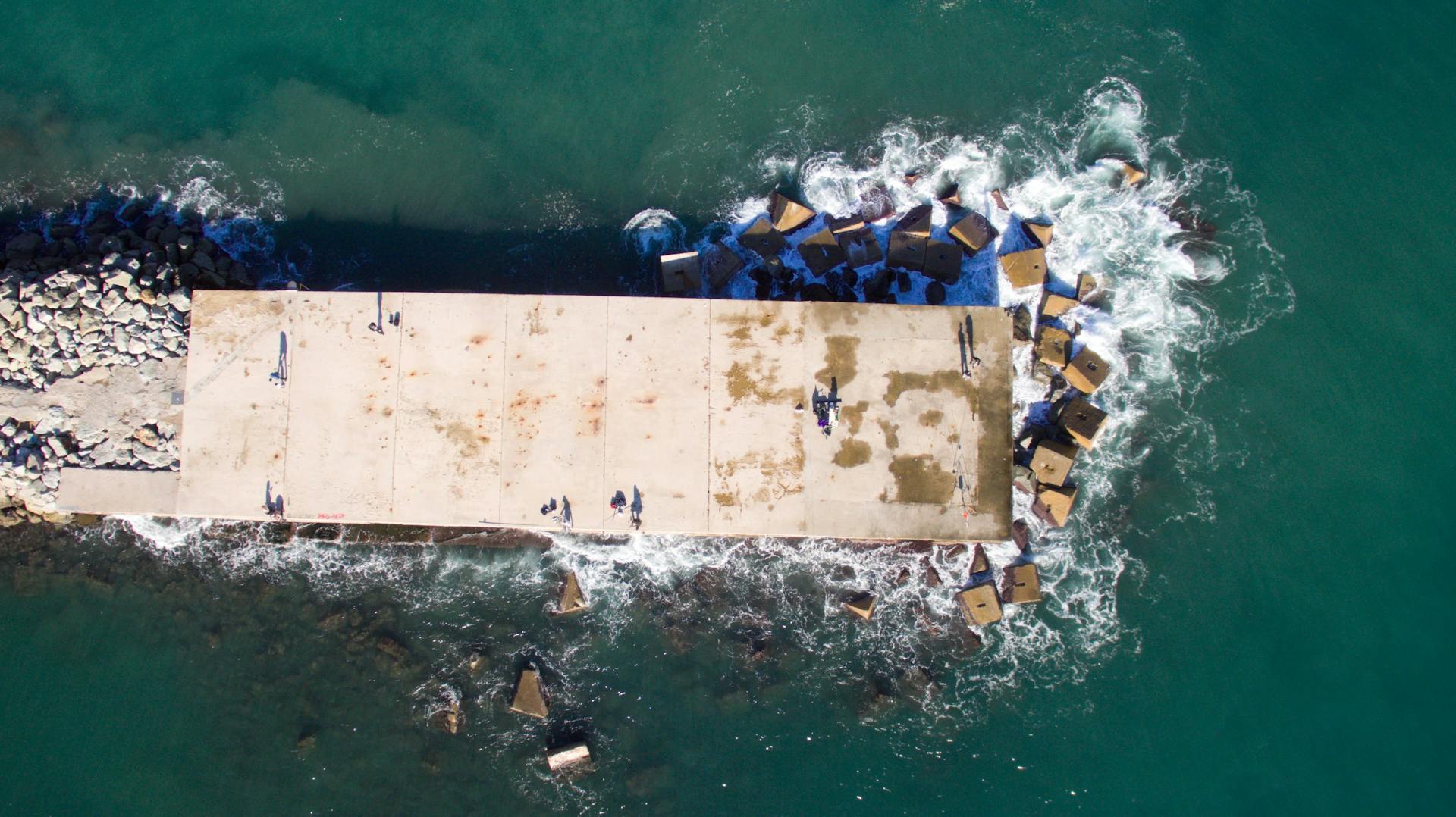 Aerial view of a concrete platform extending into the ocean with waves crashing around it.