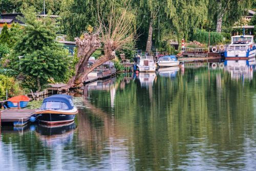 Free Peaceful riverside scene with docked boats and lush trees reflecting in calm water. Stock Photo