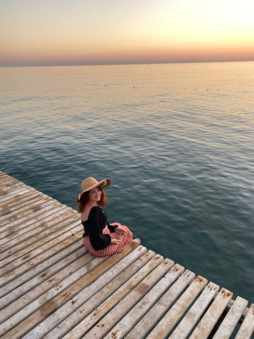 High-Angle Shot of a Woman Wearing Sun Hat while Sitting on Wooden Dock during Sunset