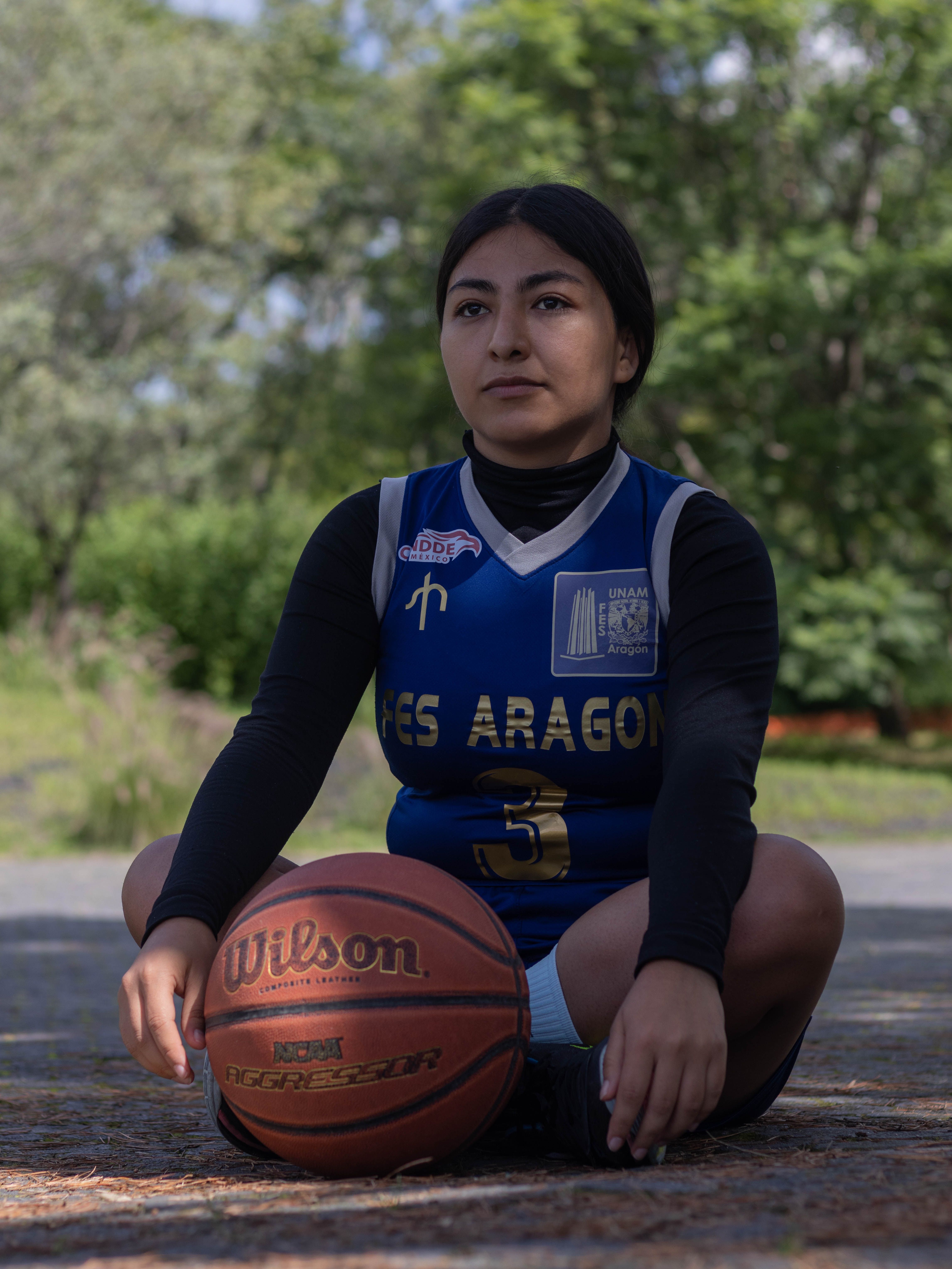 A young woman sitting outdoors in a basketball jersey, holding a basketball.