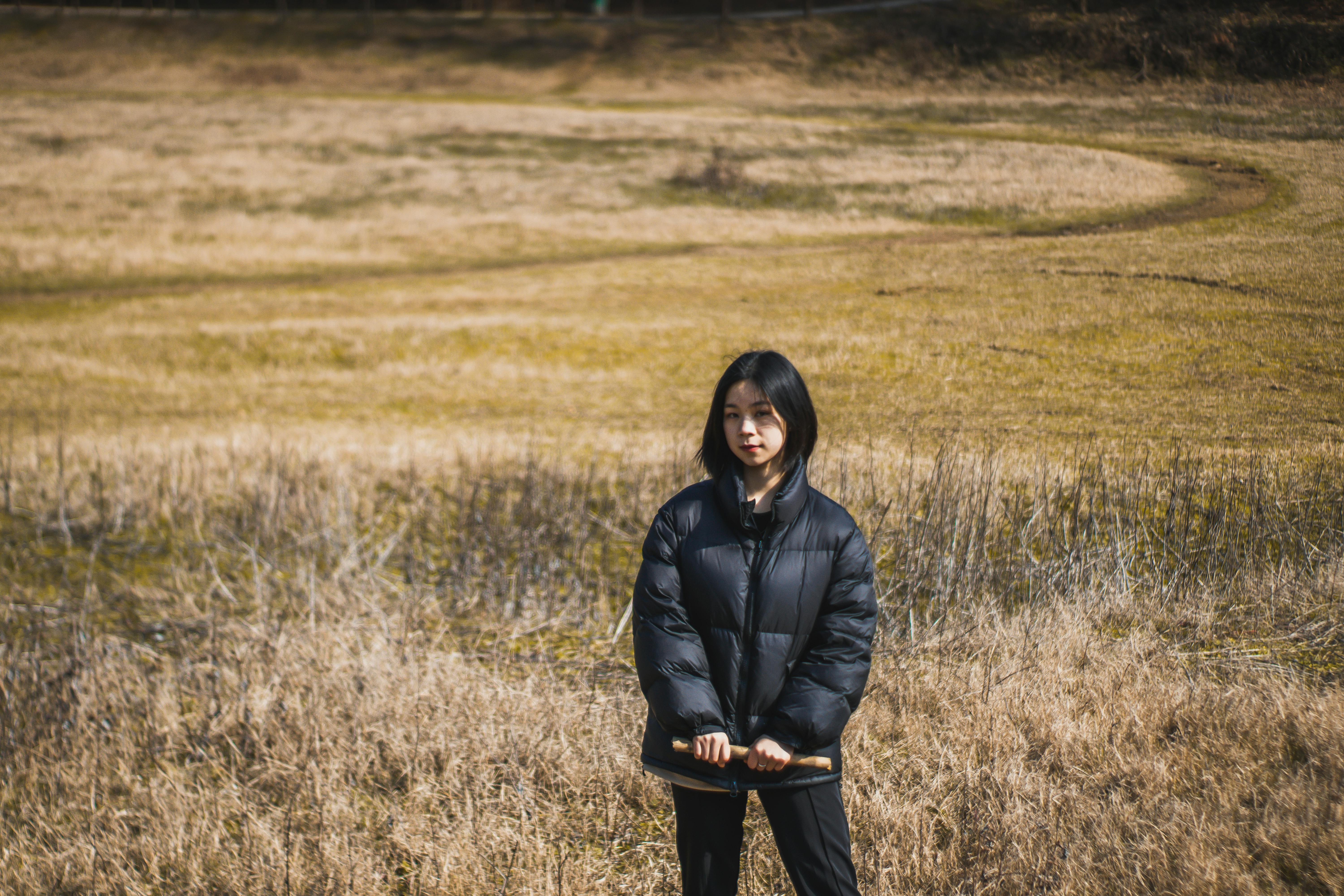 A young woman in a black jacket stands holding a stick in a grassy field during the day.