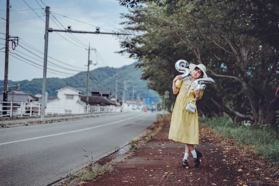 バルーンを持った黄色のドレスの女性が木々に囲まれた田舎の歩道に立っています。
