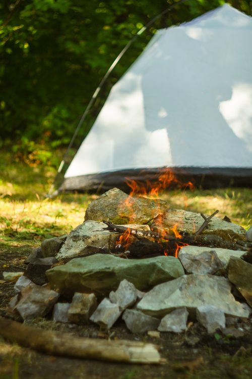 Free A warm campfire in a stone ring near a tent in a lush forest setting, ideal for camping enthusiasts. Stock Photo