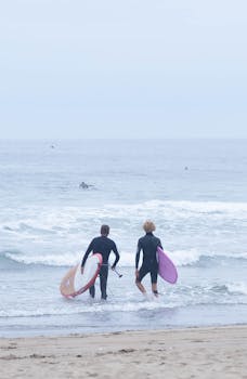 Two surfers with boards heading into the ocean on a cloudy day at Avilés Beach, Spain.