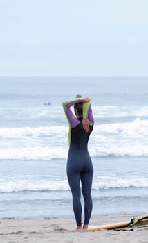 Woman in wetsuit stretching on Avilés beach with surfboard nearby. Perfect for fitness and lifestyle themes.