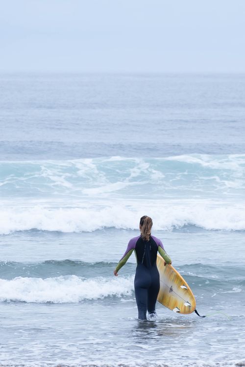 Free A woman in a wetsuit walks into the sea with a surfboard, ready to surf. Stock Photo