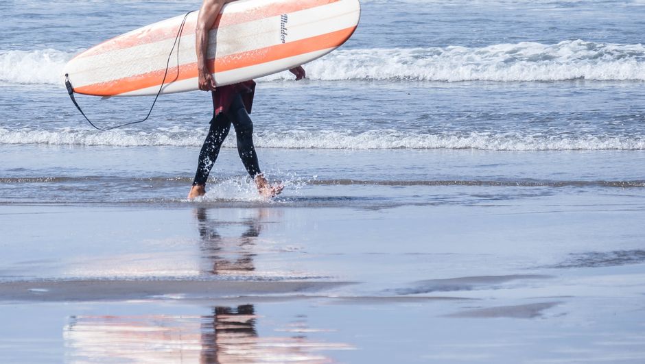 Surfer carrying board along Avilés beach in Spain, reflecting in wet sand.