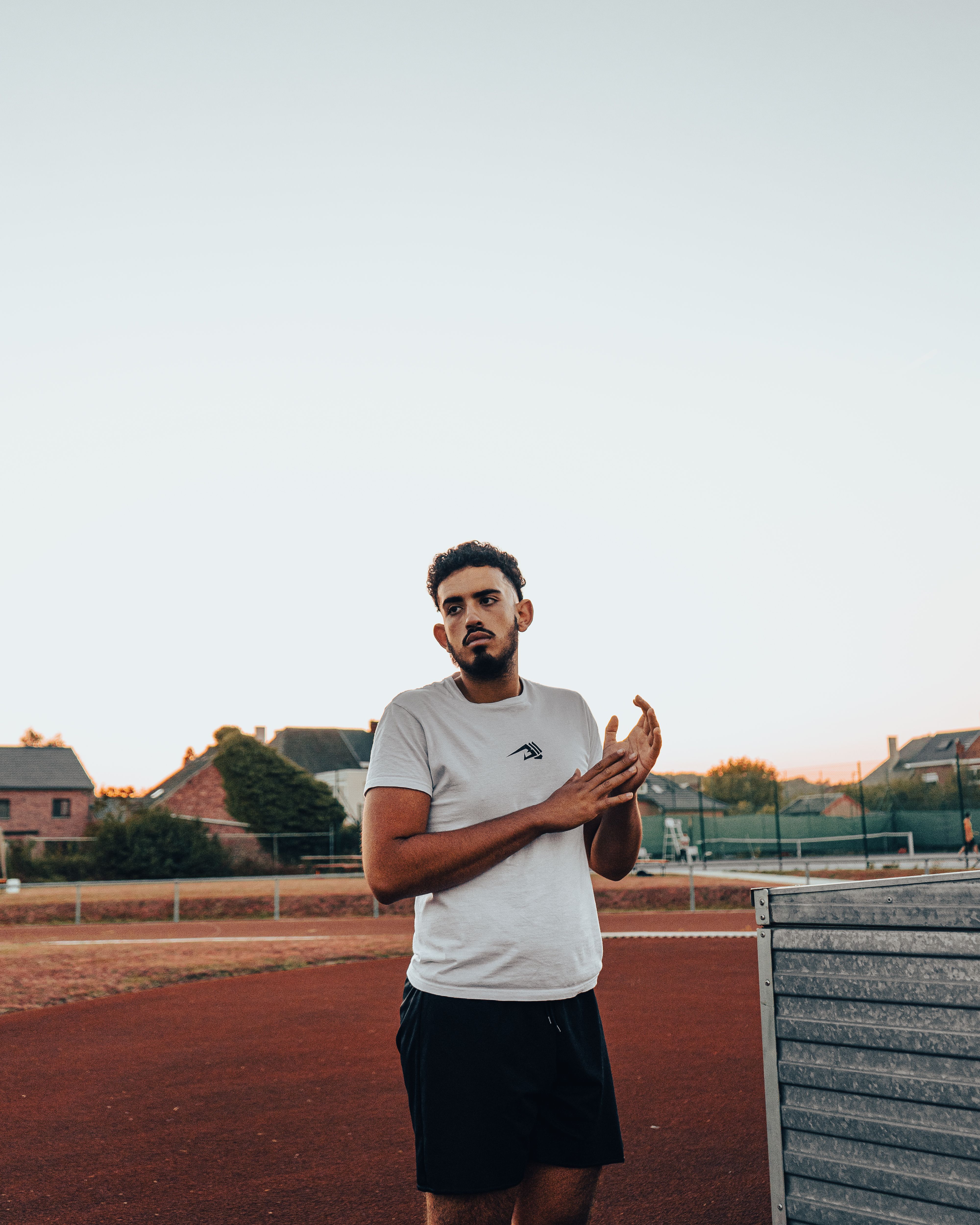 A young man stands on a sports field in Andenne, Belgium during sunset, with modern houses in the background.