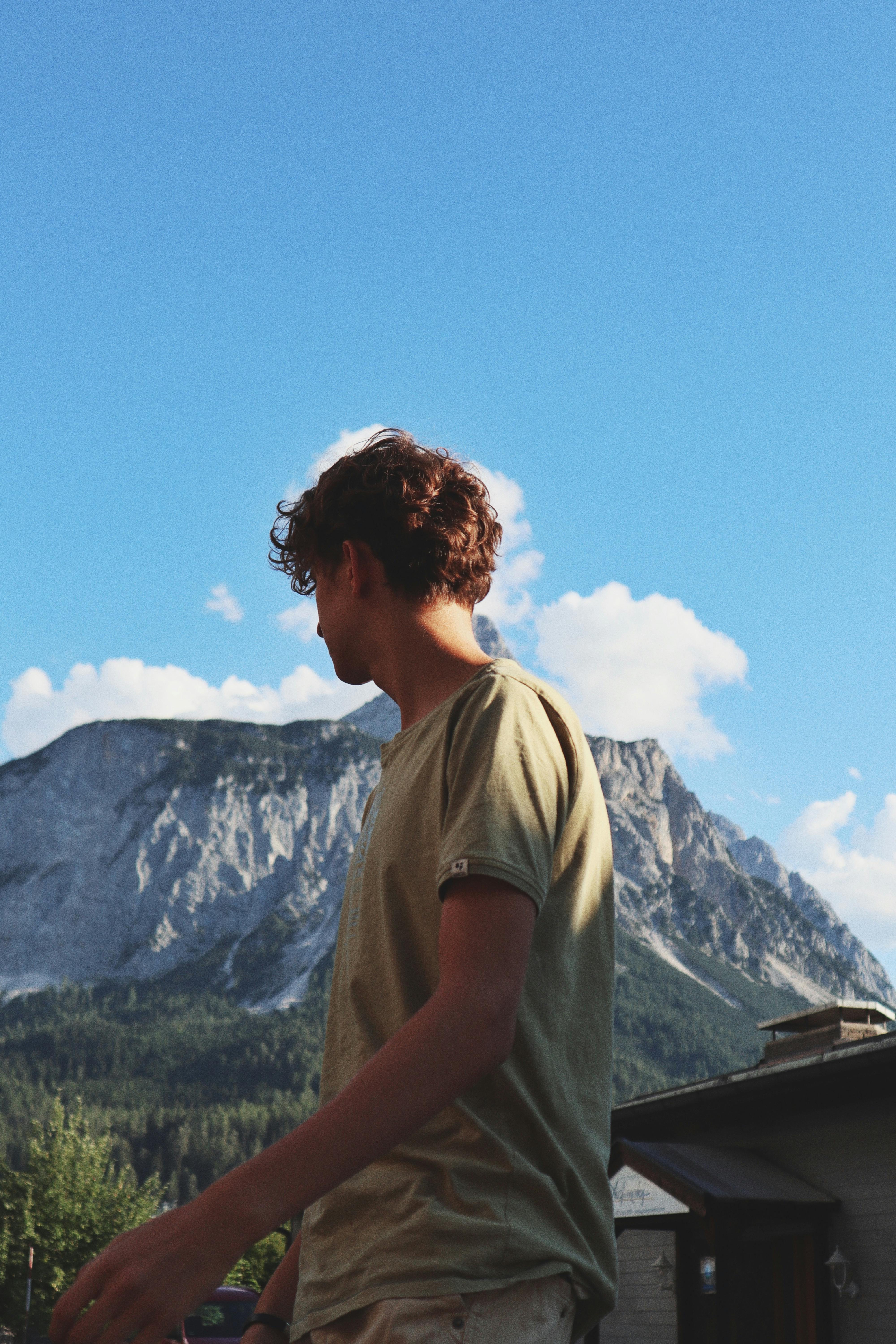 A young man stands outdoors near a mountain with a bright blue sky, embracing nature.
