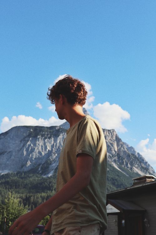 Free A young man stands outdoors near a mountain with a bright blue sky, embracing nature. Stock Photo