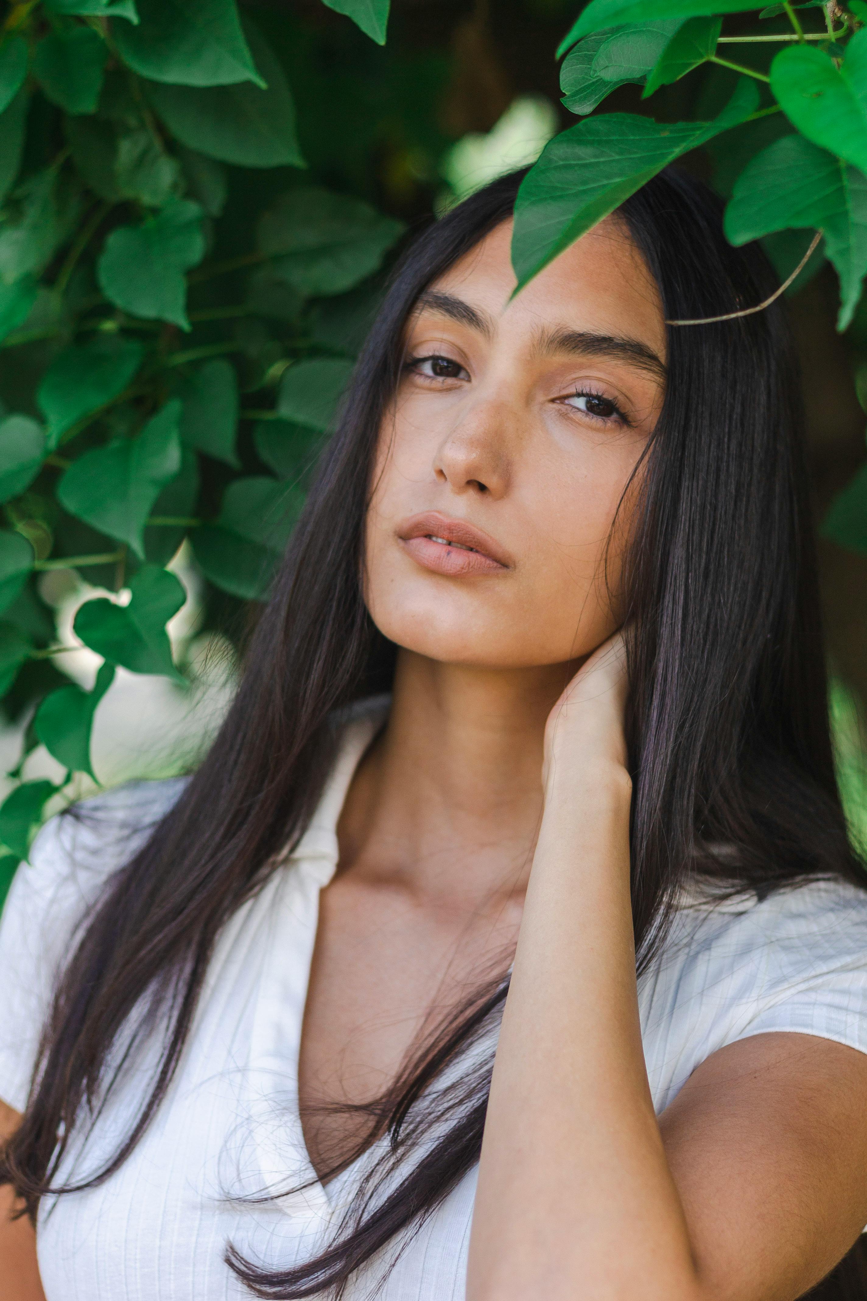 Beautiful woman with black hair poses gracefully among lush green leaves, showcasing natural elegance.