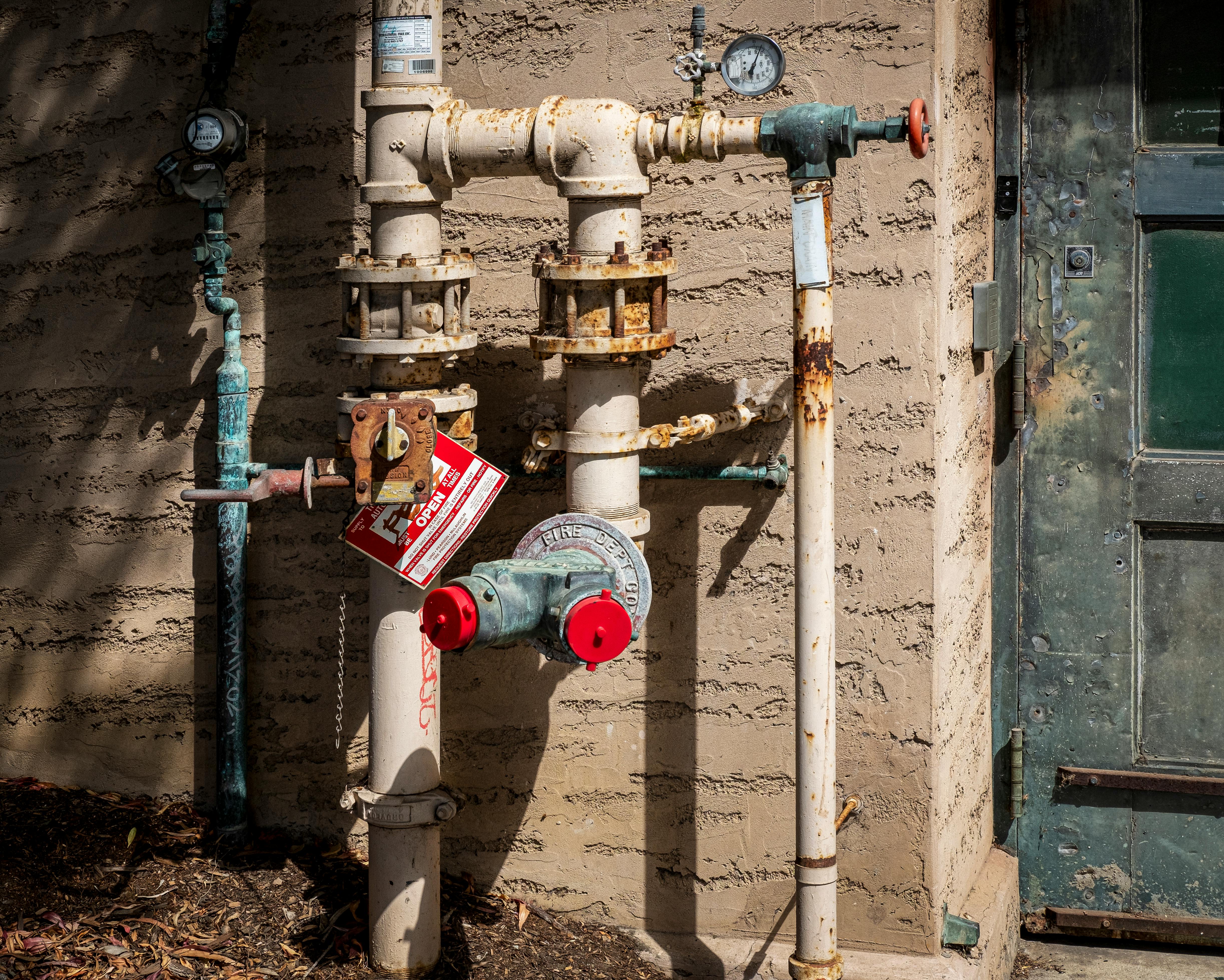 Rusty outdoor plumbing pipes with pressure gauges and warning signs on a wall.