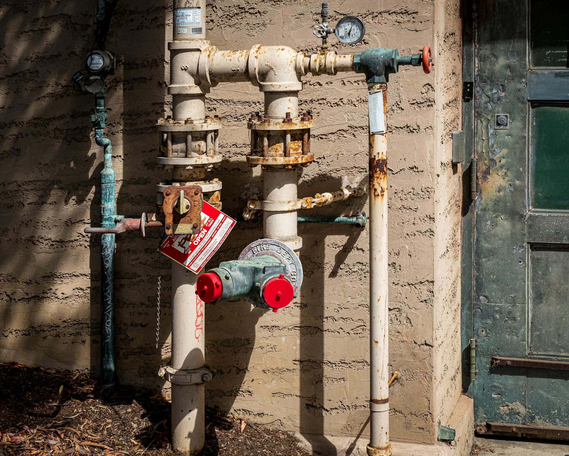 Rusty outdoor plumbing pipes with pressure gauges and warning signs on a wall.