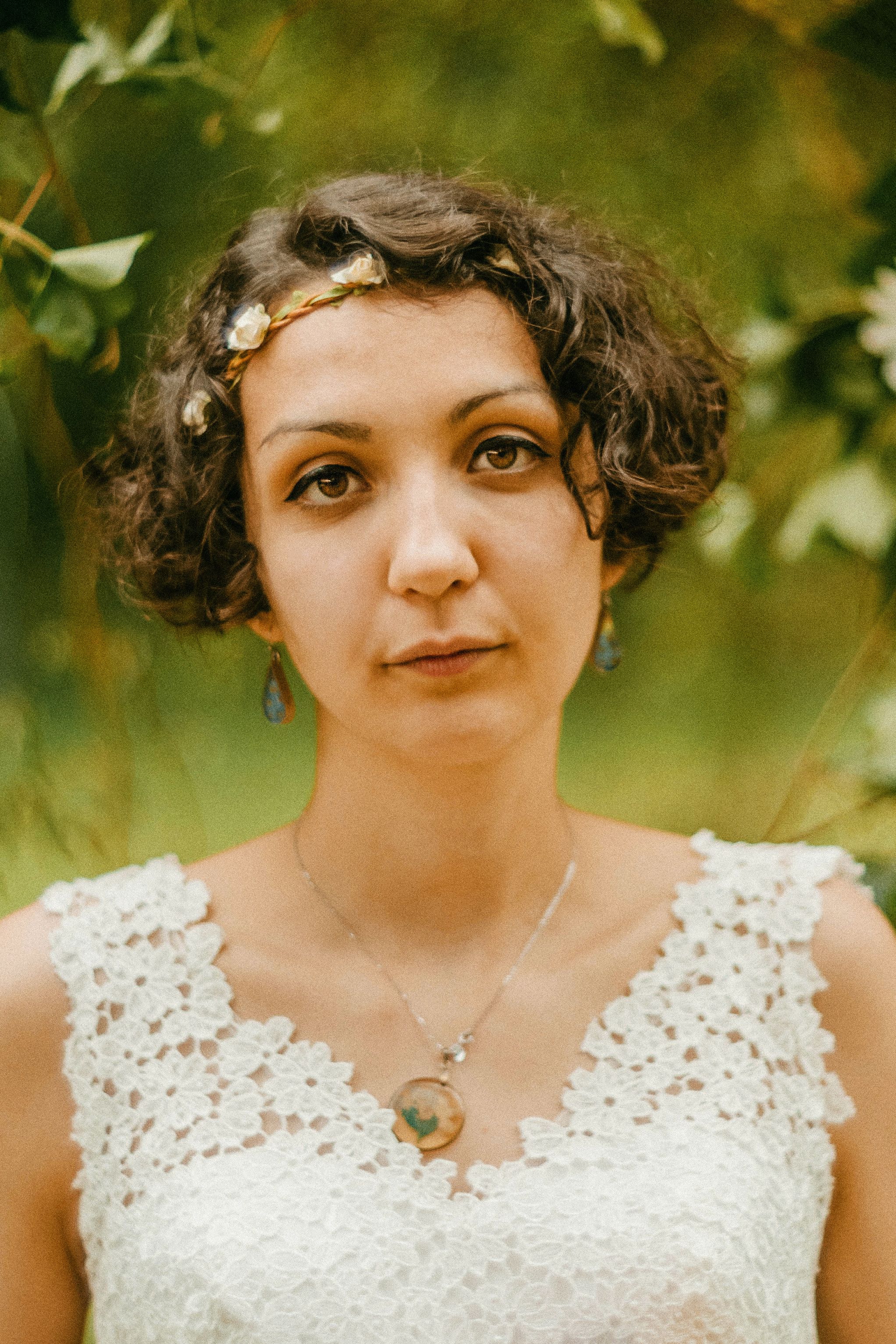 Portrait of a woman in a white lace top and floral headband, set against a natural backdrop.