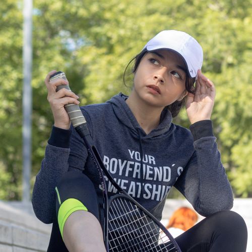 Free A young woman in casual sportswear sits outdoors holding a tennis racket, wearing a white cap and hoodie. Stock Photo