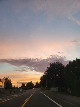 A tranquil road scene at sunset with a picturesque sky filled with pastel clouds and trees lining the street.