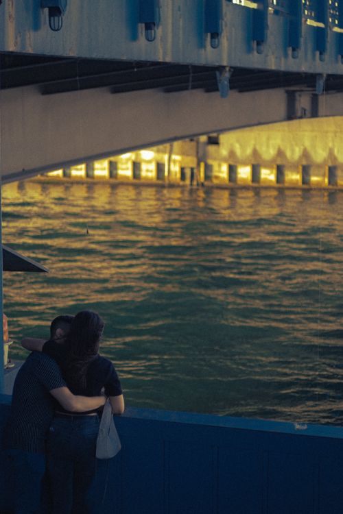 Free A couple embracing beneath an urban bridge by the river, creating a romantic scene. Stock Photo