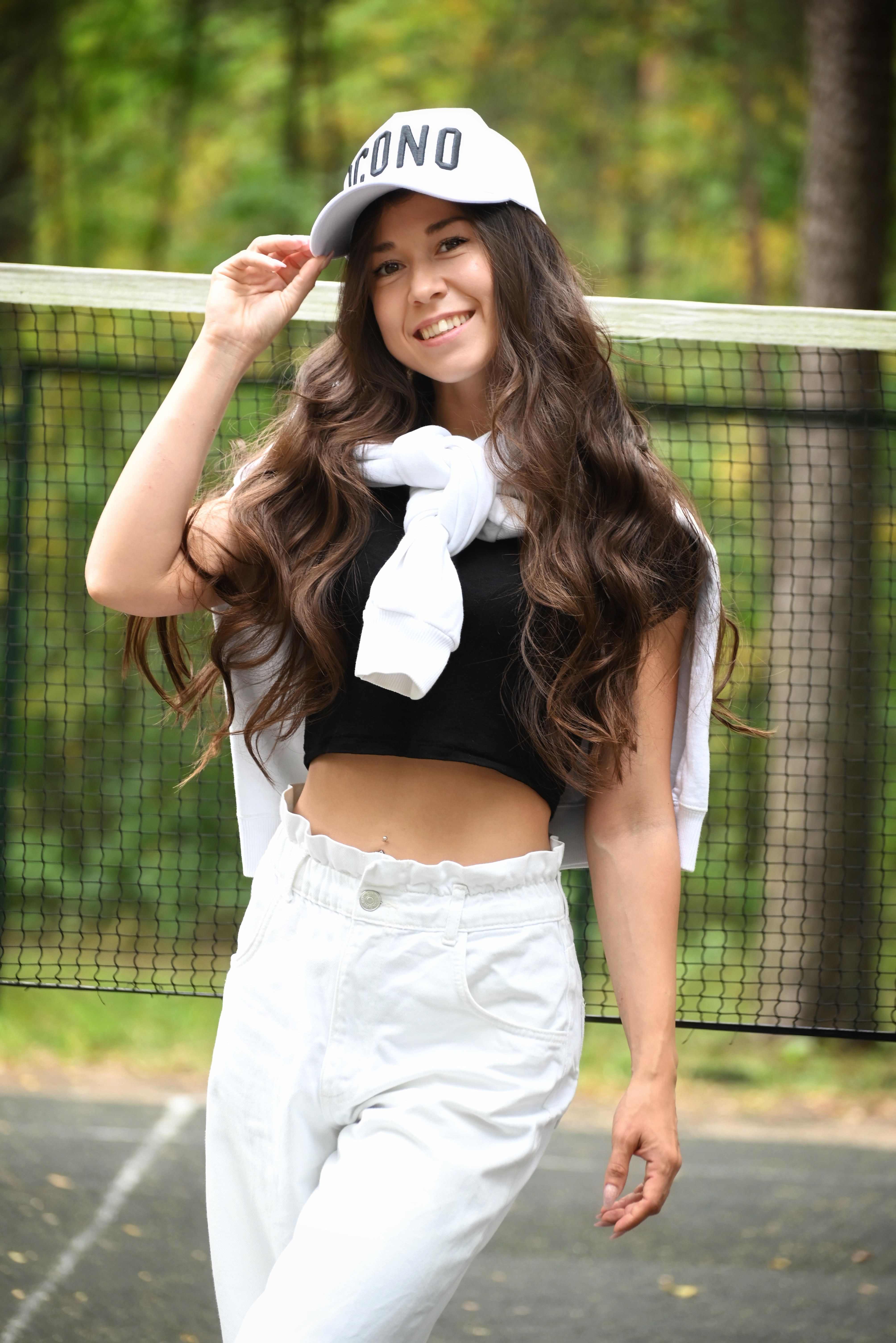 Fashionable young woman in crop top and cap posing on tennis court outdoors.