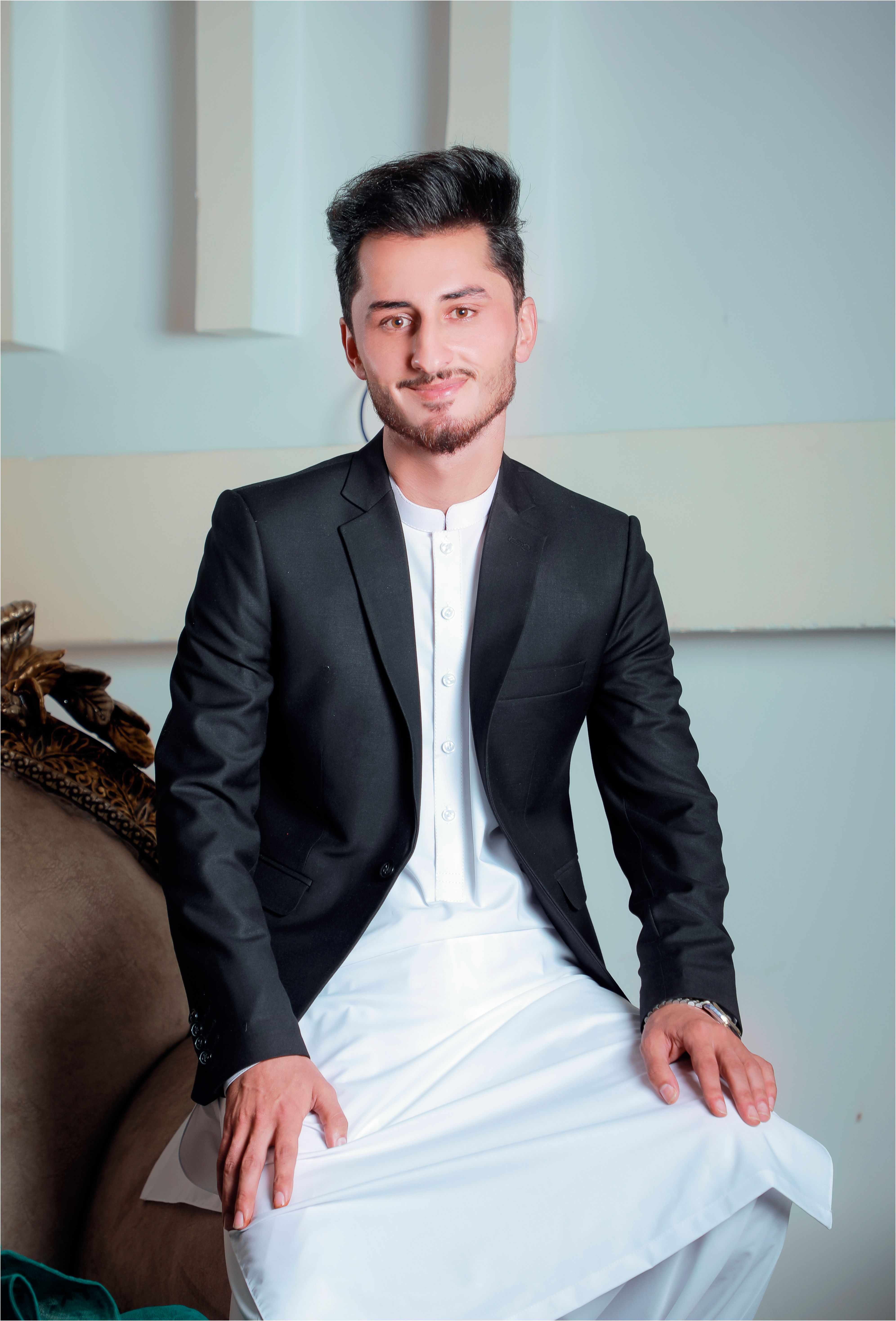 Stylish young man in a black suit and traditional dress, smiling confidently indoors.
