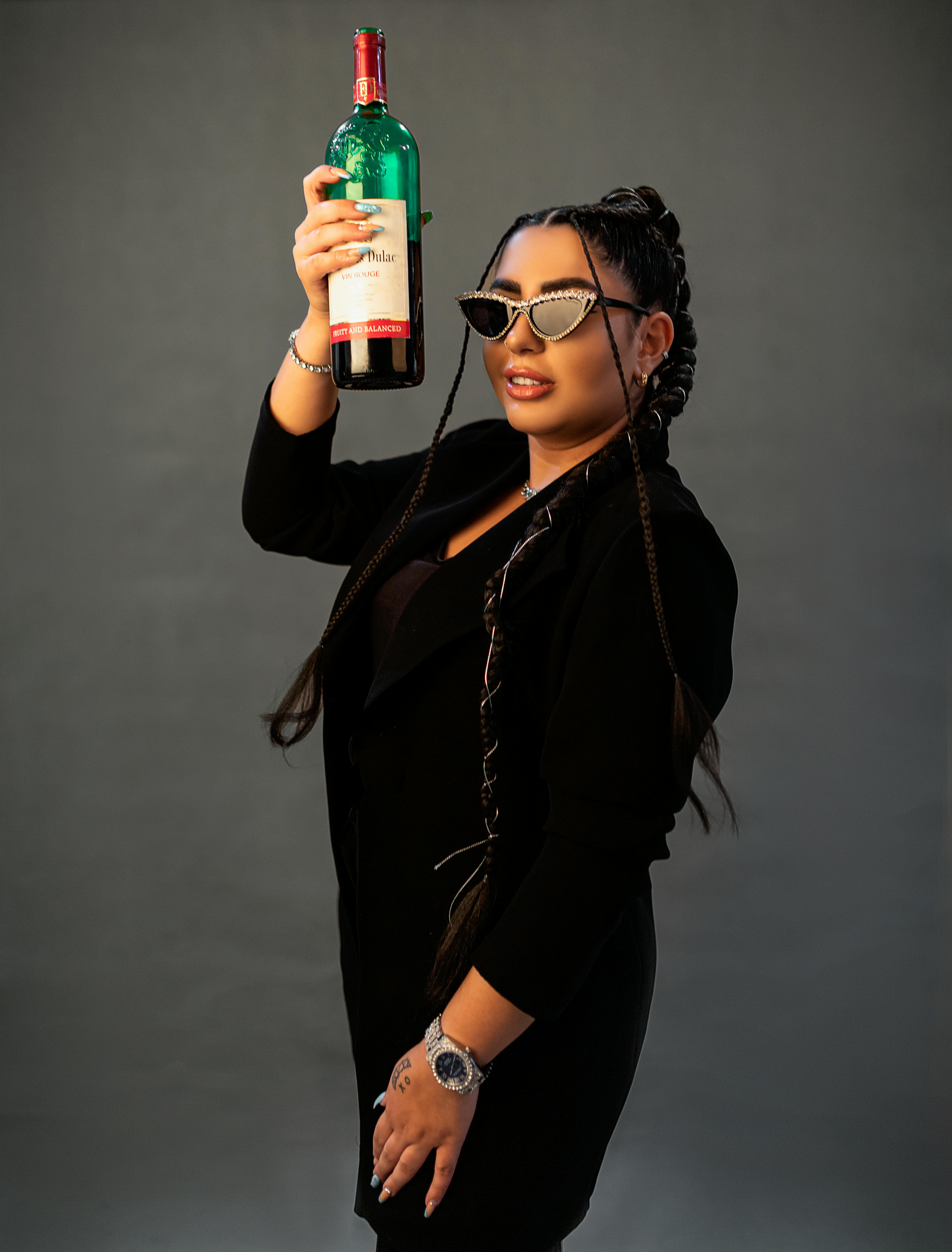 Fashionable woman in black outfit with braids holding a wine bottle against a gray background.