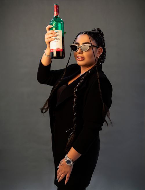 Free Fashionable woman in black outfit with braids holding a wine bottle against a gray background. Stock Photo