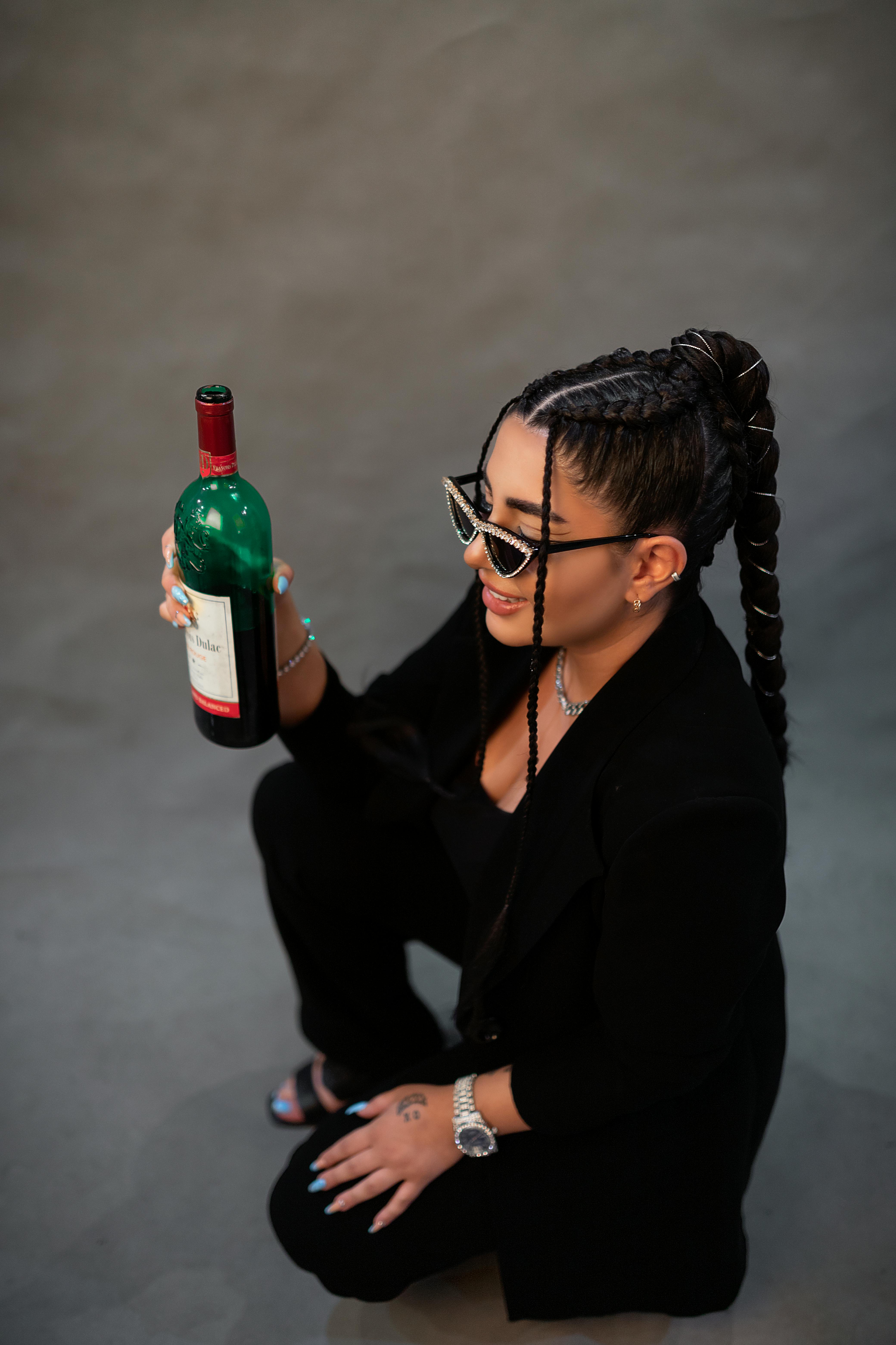 Stylish woman in black with braids holds red wine, showcasing modern fashion in Tehran studio.