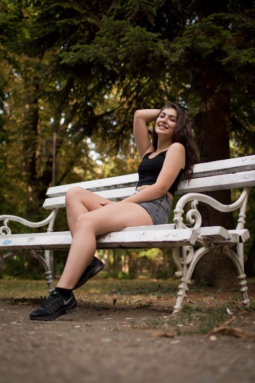 Free A cheerful woman sitting on a white bench in a sunny park, expressing joy and relaxation. Stock Photo