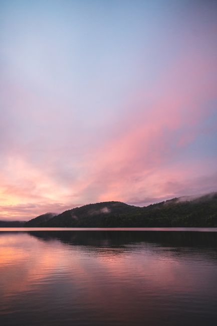 Beautiful sunset over a tranquil lake, reflecting a purple and pink sky with silhouetted mountains.