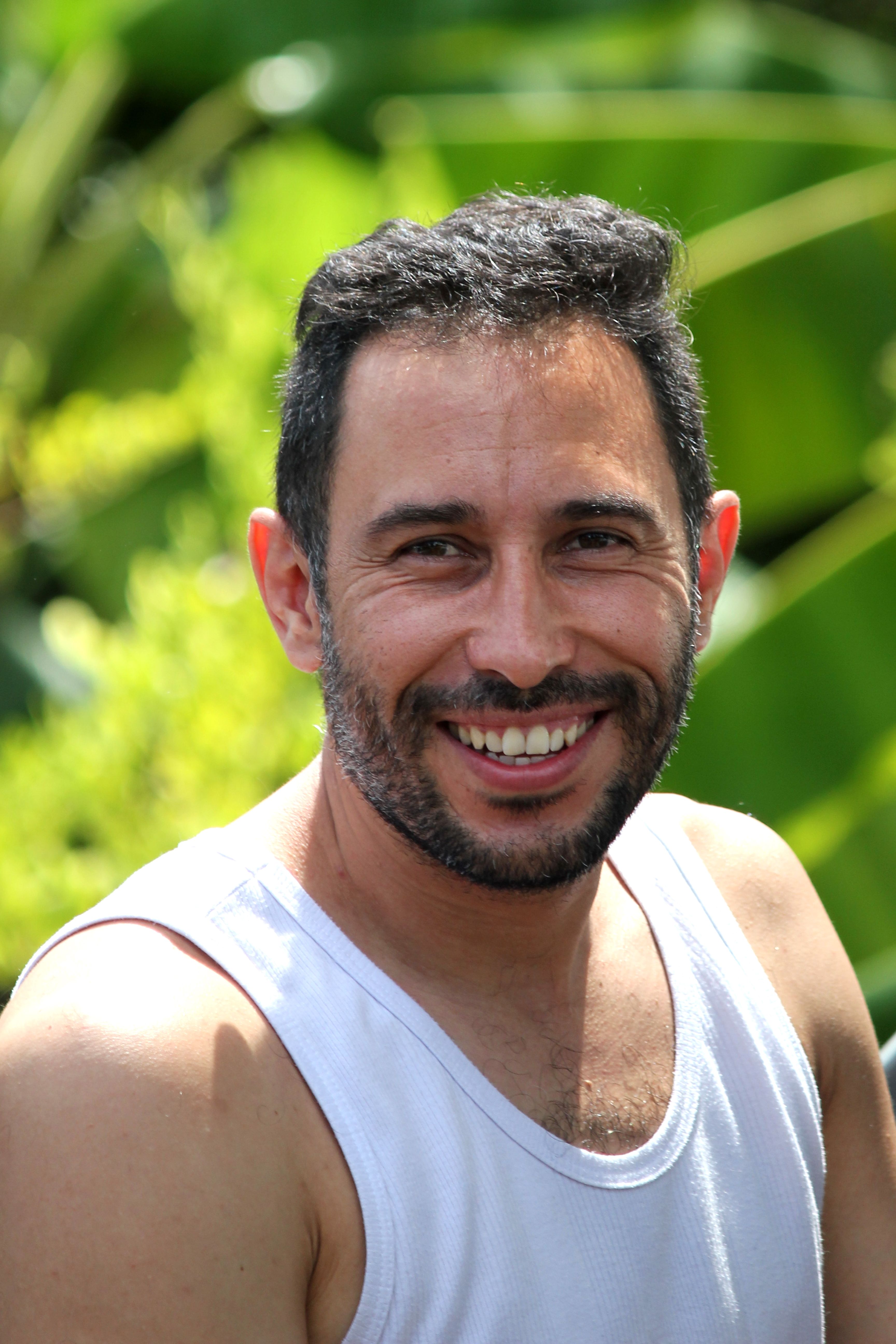 Portrait of a smiling man in a white tank top against lush green foliage, emphasizing natural joy.