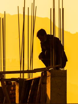 A silhouette of a man sitting at a construction site during a golden hour sunset, with a warm orange background.