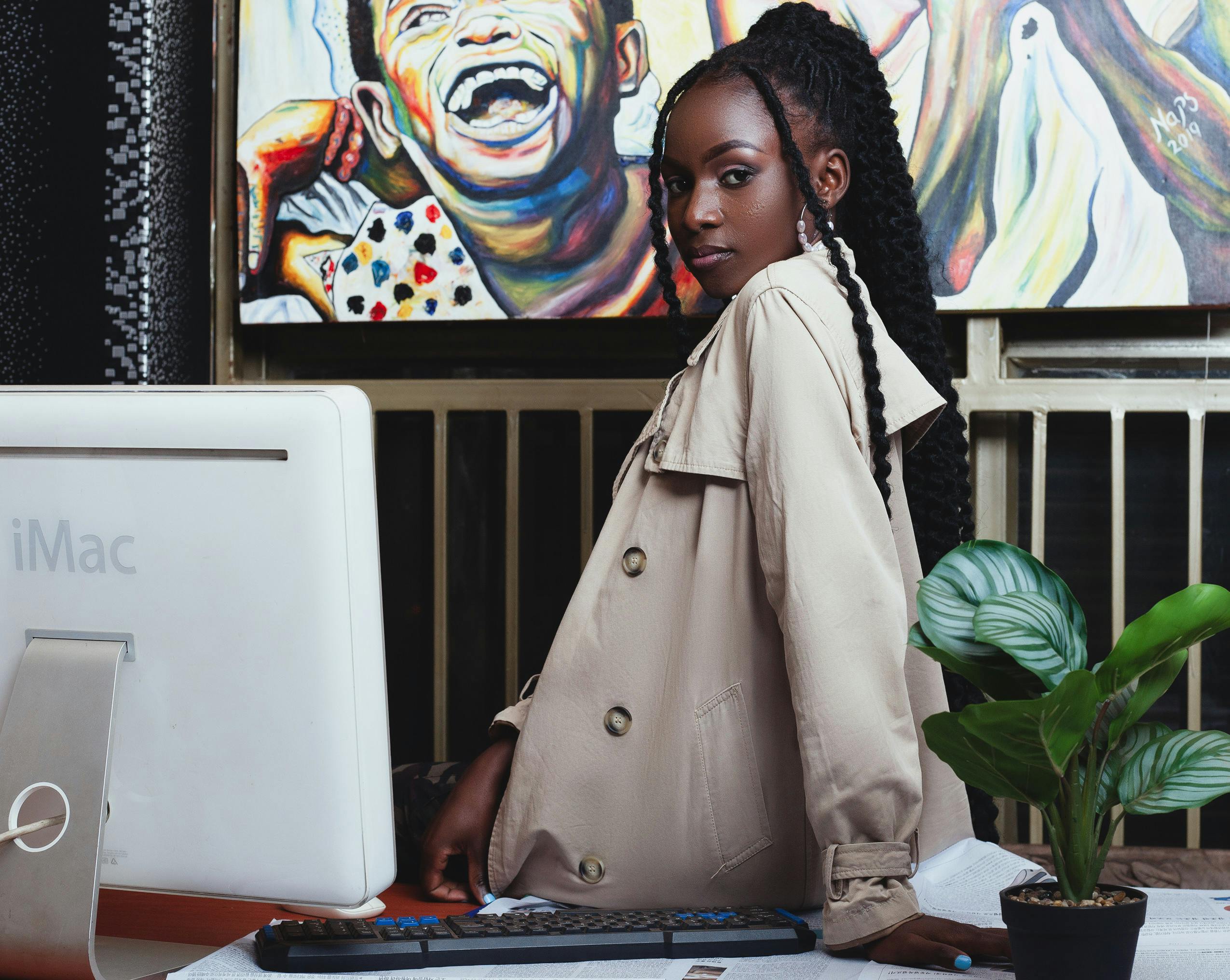 African woman in a beige coat poses confidently indoors by a desk in Dodoma. Artistic background.