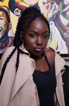 A confident woman in a beige coat and braided hair poses against a colorful art backdrop.