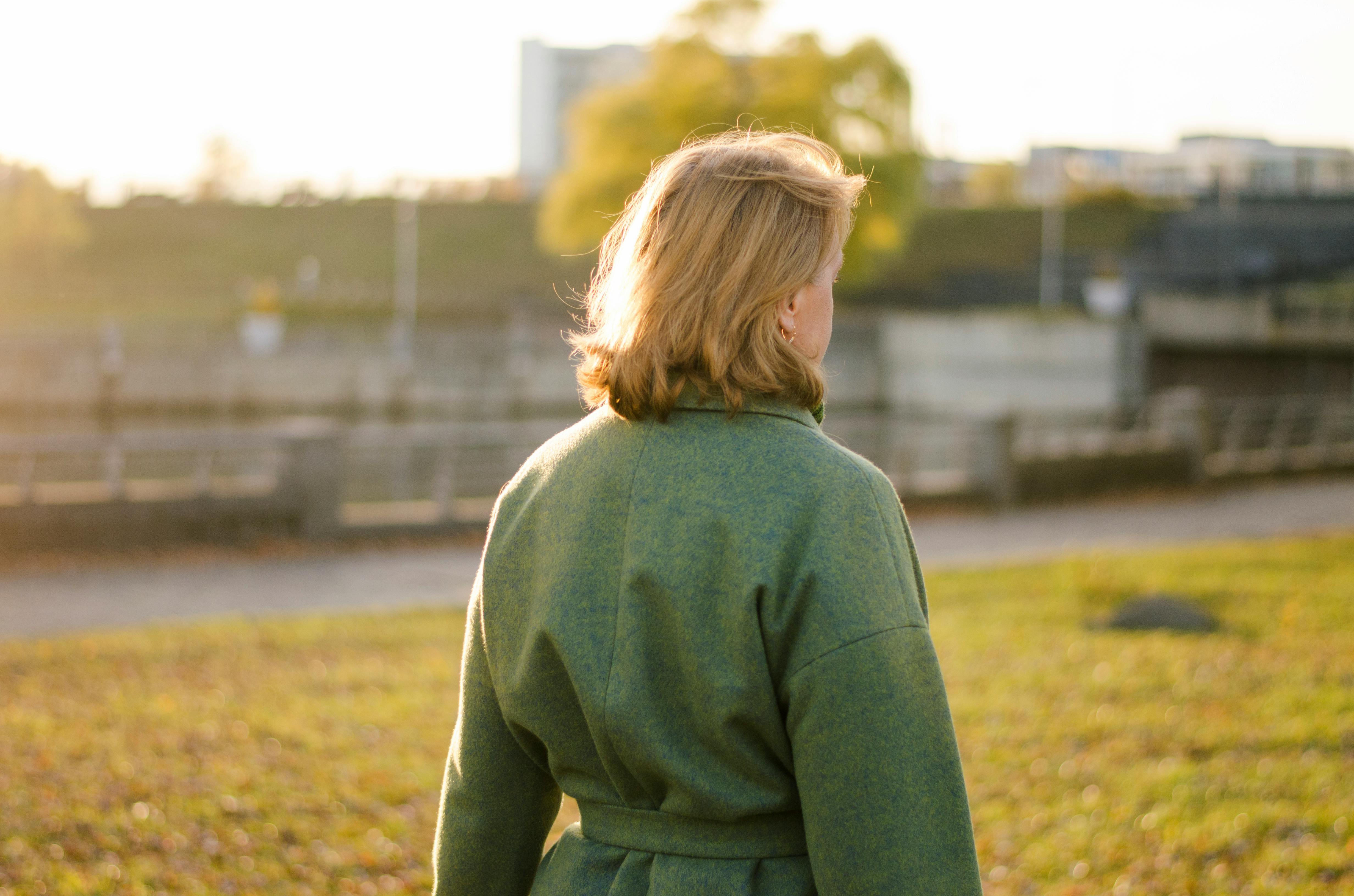 Rear view of a woman in a green coat, basking in sunlight outdoors, capturing tranquility and leisure.