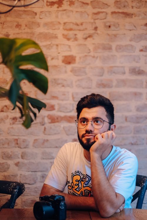 Free Man with glasses and beard thoughtfully looking up while seated indoors, camera on table. Stock Photo
