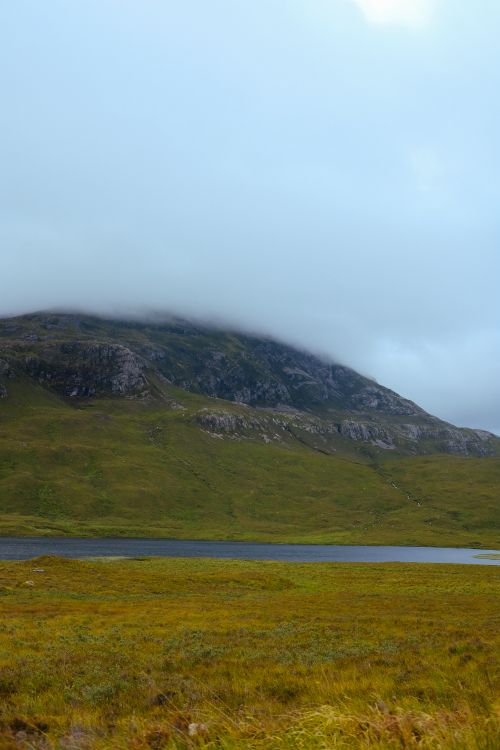 Free Serene landscape of a cloud-covered mountain, lush green fields, and a calm lake. Stock Photo