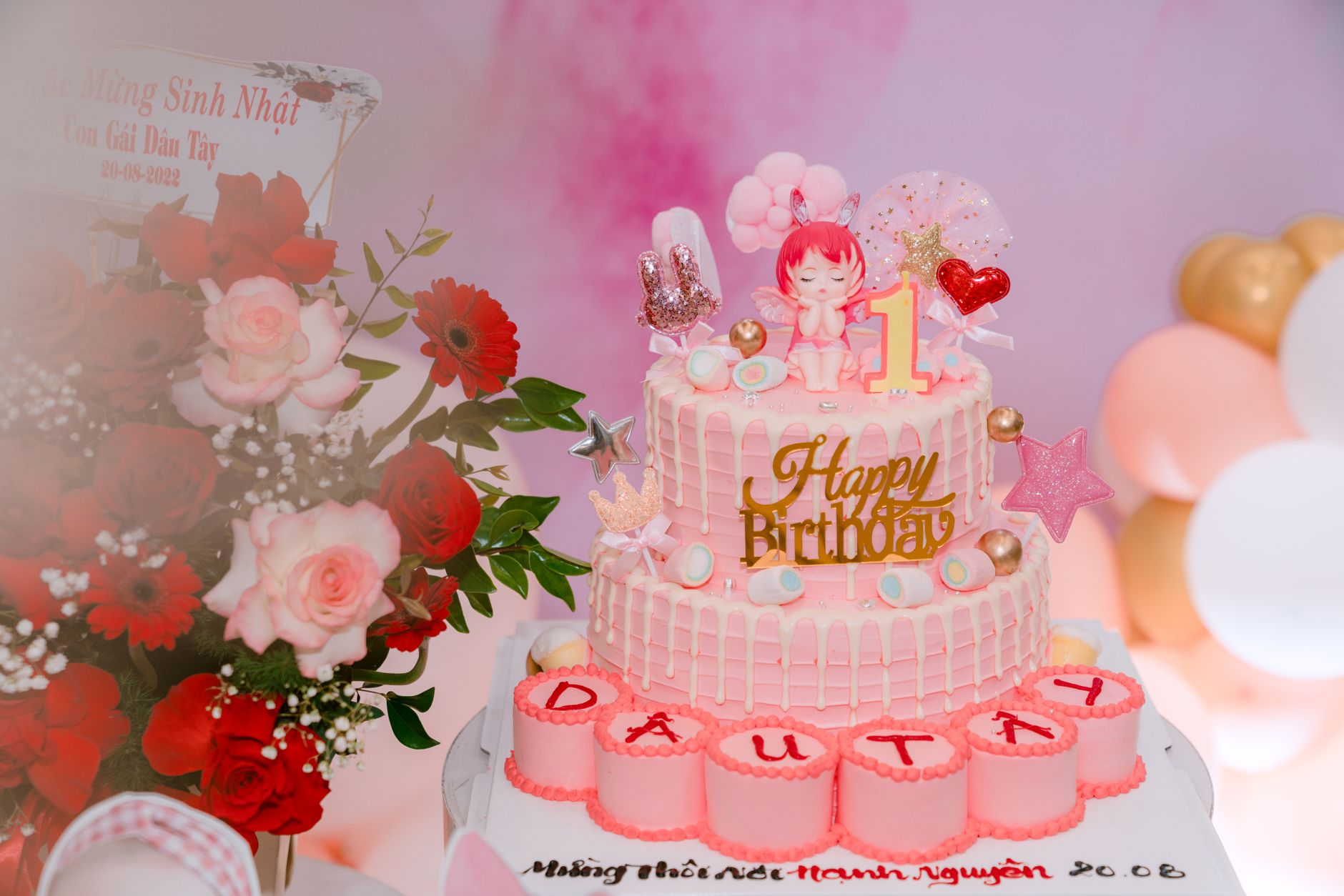 Adorable 1st birthday cake with pink decorations, toys, and floral backdrop.