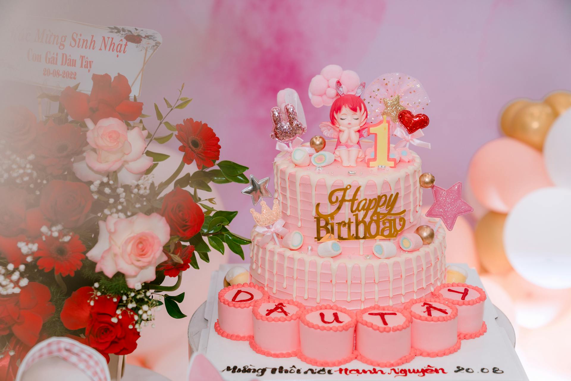 Adorable 1st birthday cake with pink decorations, toys, and floral backdrop.