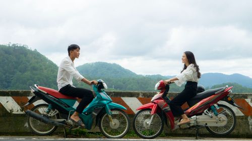Free A man and woman sit on motorbikes facing each other on a scenic outdoor road with mountains. Stock Photo